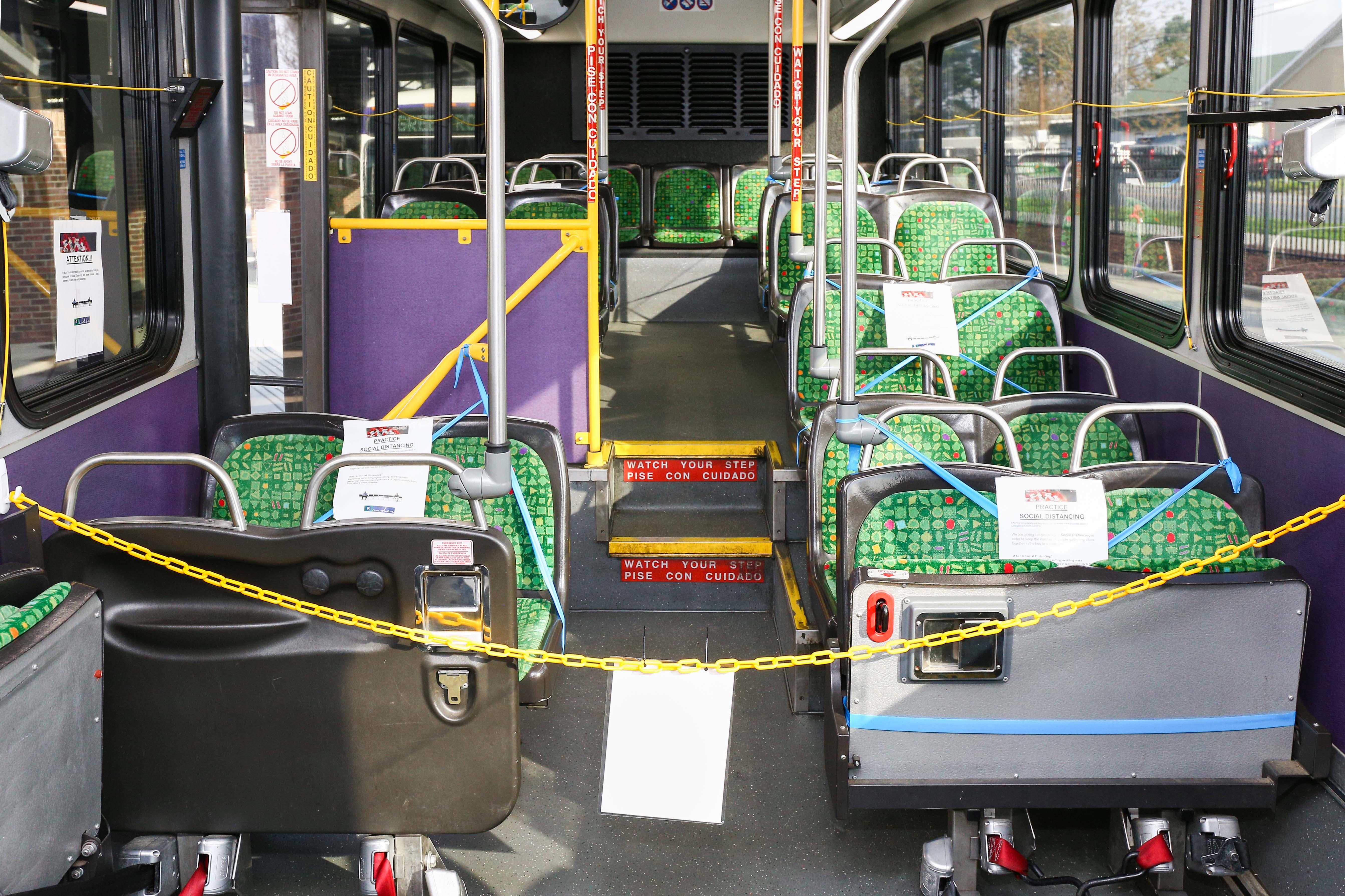 Picture of interior of a bus with yellow chain to show that front of bus is closed.  Seats are marked off with a sign for passengers to not sit in those seats to increase social distancing. 